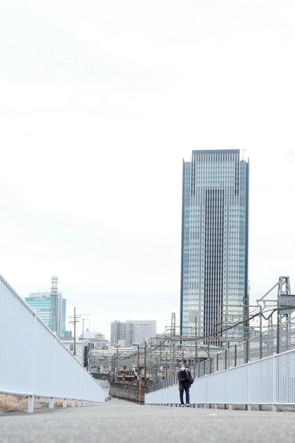 An elderly lady strolling across the bridge, with the towering structures of Nagoya Station in the background.