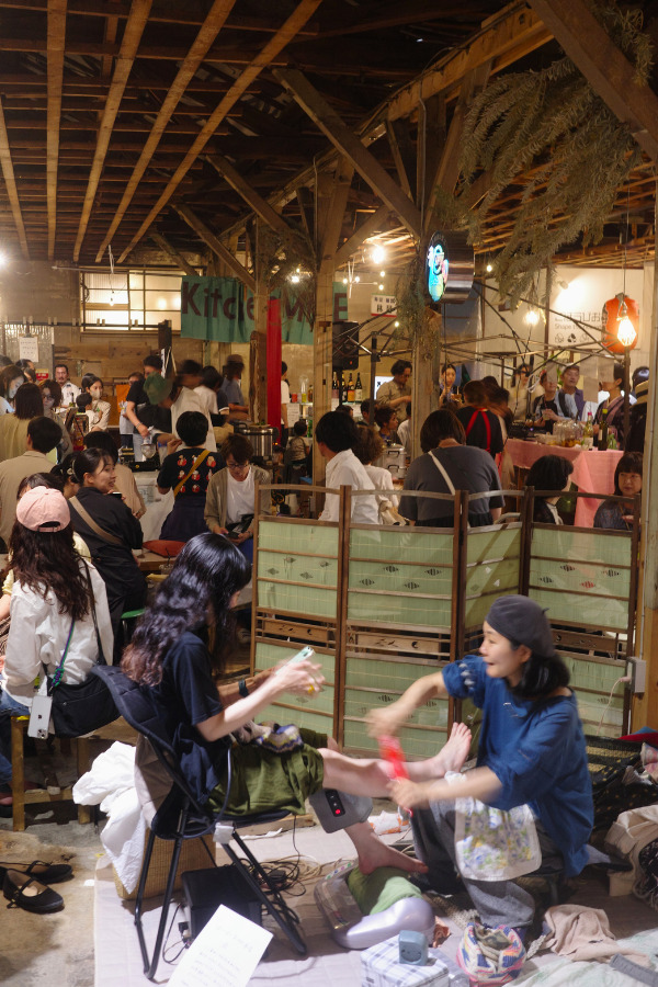 Inside Kinjo Market at night, bustling with people enjoying various activities, including a massage session in the foreground.