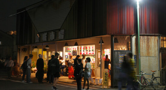 People gathering outside Kinjo Market at night, with the market brightly lit and vibrant decorations visible through the windows