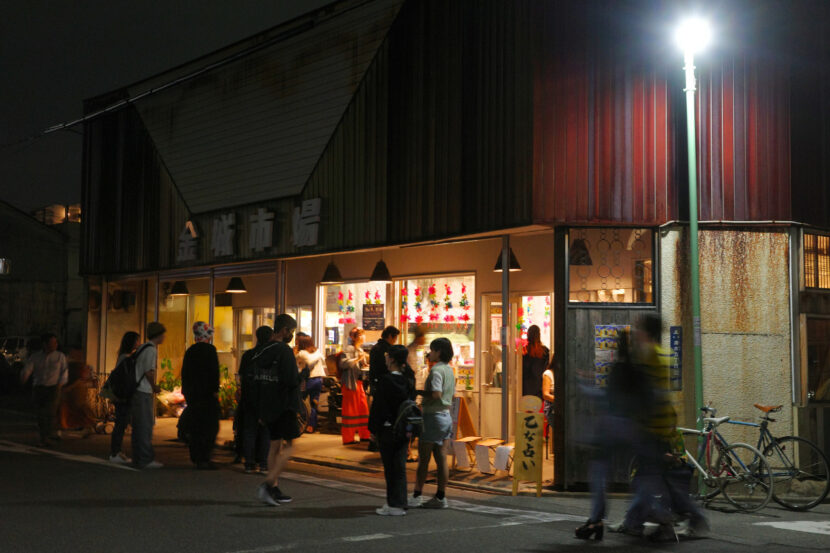 People gathering outside Kinjo Market at night, with the market brightly lit and vibrant decorations visible through the windows