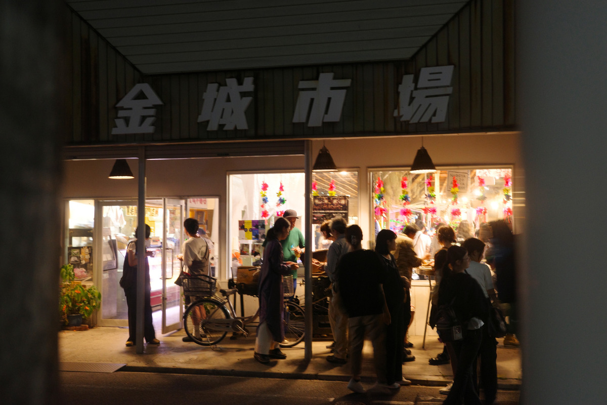 People gathered outside Kinjo Market at night, with the market's bright lights and decorations visible through the windows