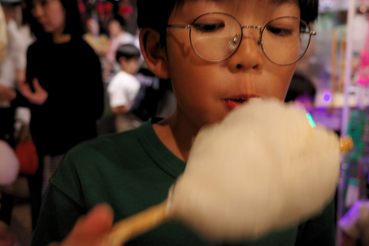 A child enjoying cotton candy at Kinjo Market, with other visitors and vibrant decorations in the background.