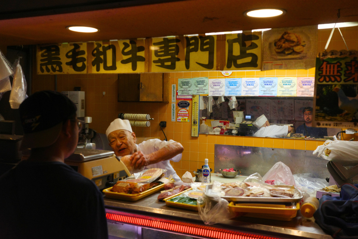 An elderly vendor serving customers at a stall specializing in Kuroge Wagyu beef inside Kinjo Market.